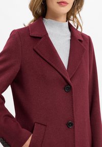 Cappotto bordeaux in lana con rever a scanalatura, due tasche frontali e bottoni neri, abbinato a un dolcevita grigio a coste sottostante.