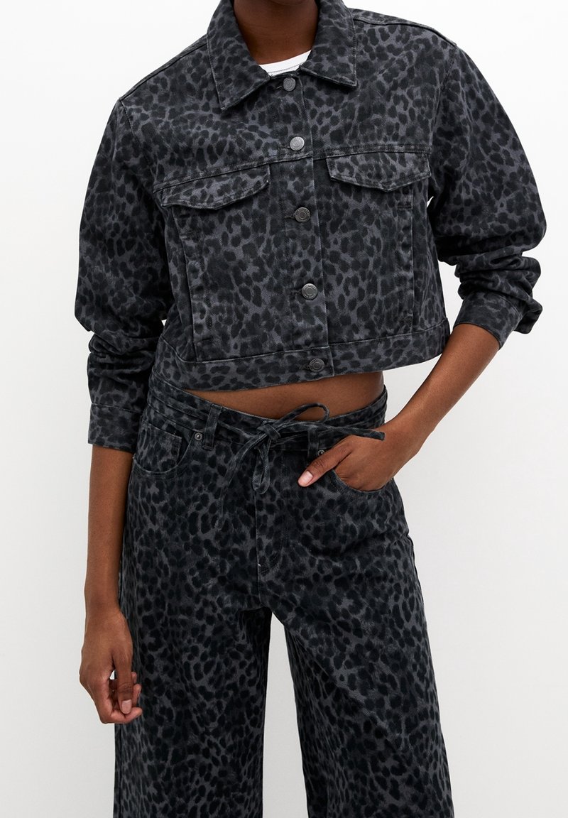 Veste en jean noir raccourcie et pantalon taille haute assorti avec un subtil imprimé léopard foncé, porté par une personne avec une main dans la poche.