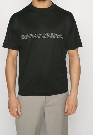 Mężczyzna w czarnej koszulce Emporio Armani z białym logo i jasnoszarych spodniach, stojący na jednolitym tle.