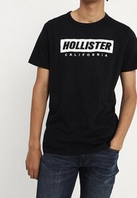 Černé bavlněné tričko s bílým potiskem, který zní "HOLLISTER CALIFORNIA." Kulatý výstřih, krátké rukávy, volný střih.