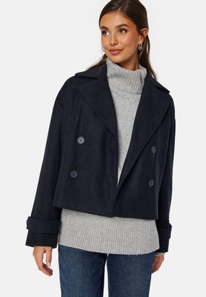 Manteau bleu marine à double boutonnage, en mélange de laine douce, avec de grands revers et des accents de boutons, superposé à un pull gris à côtes.
