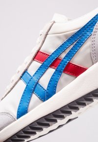 Sneakers con tomaia in tela bianca, righe blu e rosse, dettagli in suede grigio, suola in gomma nera testurizzata e cuciture visibili.