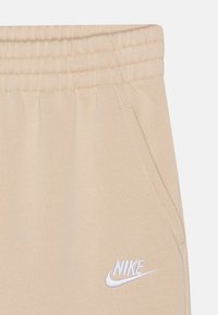 Bežinės kelnės su elastingu juosmeniu, turinčios šonines kišenes ir baltą „Nike“ logotipą išsiuvinėtą ant apatinės kojos. Minkšta audinio tekstūra.