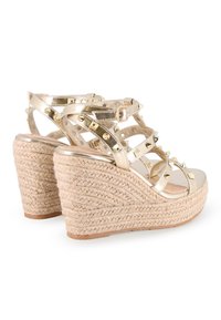 Sandalias cuña metálicas doradas con tacones de paja texturizada. Cuenta con acentos de tachuelas y correas entrecruzadas. Cierre ajustable con hebilla en el tobillo.