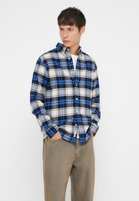 Camisa de flanela xadrez azul e cinza com colarinho abotoado, mangas longas e um ajuste relaxado, combinada com calças bejes.