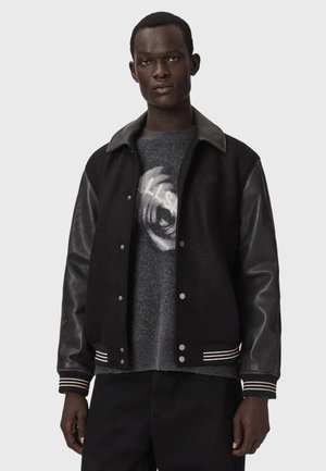 Homme portant une veste varsity noire avec des manches en cuir par-dessus un pull gris avec un motif circulaire blanc, debout devant un fond uni.
