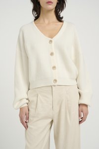 Krémový rebrovaný cardigan s V-krkom, s piatimi prednými gombíkmi a spustenými plecami. Spárované s svetlo béžovými plisovanými nohavicami.