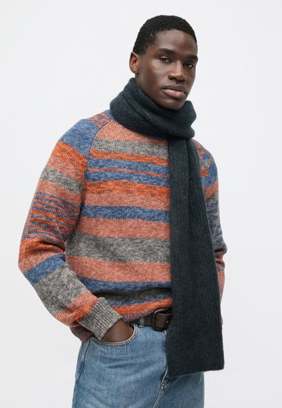 Pruhovaný vlněný svetr v oranžové, šedé a modré barvě s tmavým pleteným šátkem. Jedinec pózuje s rukama v kapsách, má ležérní vzhled.