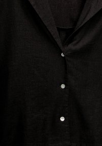 Chemise en lin noire avec col, dotée de cinq boutons blancs sur le devant. Le tissu présente une finition texturée subtile.