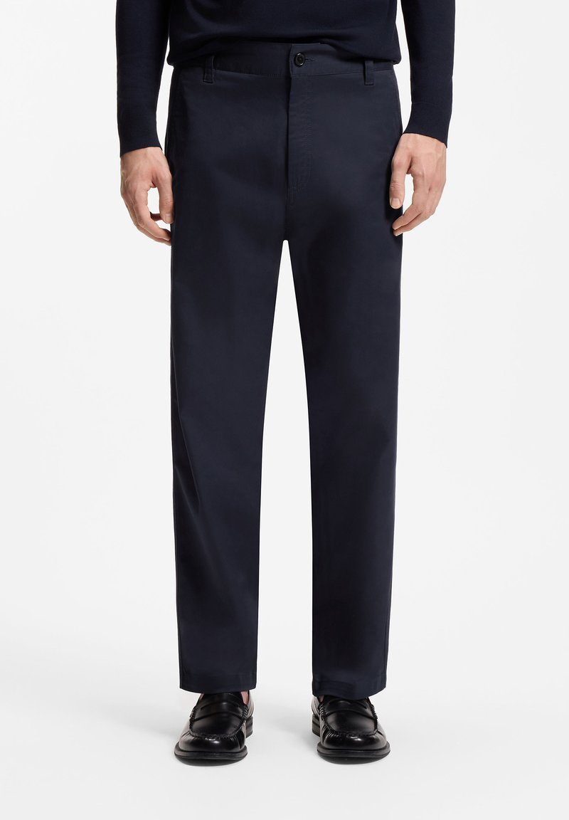 Homme portant un pantalon noir droit, une ceinture noire, un haut à manches longues foncé et des mocassins en cuir noirs, debout devant un fond blanc.