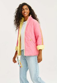 Prošívaná bunda v růžové barvě s žlutou podšívkou a manžetami. Obsahuje boční kapsy a barevný zip s třásněmi. Nesená přes bílé tričko.