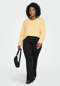 Kollane lühike kampsun, millel on kaelusel neli nööpi, kombineeritud mustade retuuside ja mustade madalate kingadega. Modell kannab väikest kotti.