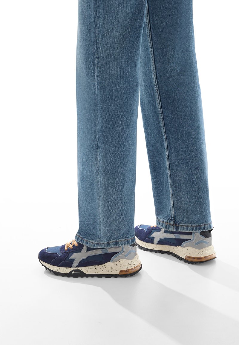 Sneakers gjorda av blå mocka och mesh med grå detaljer, spräcklig vit sula och brun häl. Kombinerat med ljusblå raka jeans.