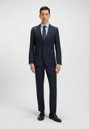 Abito blu navy con vestibilità su misura, un bottone singolo, revers a V, abbinato a una camicia azzurra e una cravatta con motivo, completato da scarpe nere.