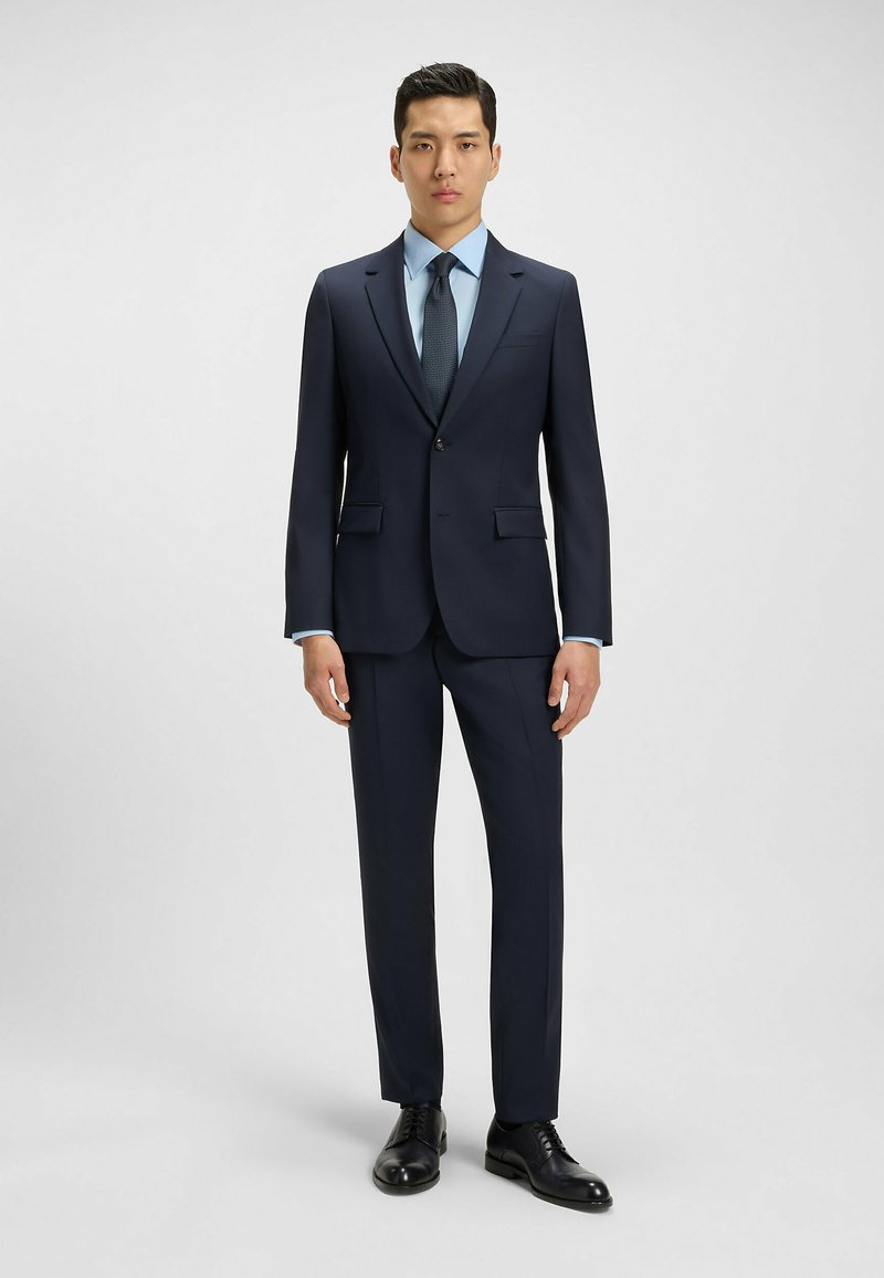 Abito blu navy con vestibilità su misura, un bottone singolo, revers a V, abbinato a una camicia azzurra e una cravatta con motivo, completato da scarpe nere.