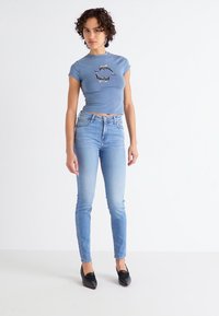 Un crop top bleu ciel avec un graphique de dauphin, associé à un jean skinny bleu clair. Mannequin portant des ballerines noires, debout contre un arrière-plan blanc.