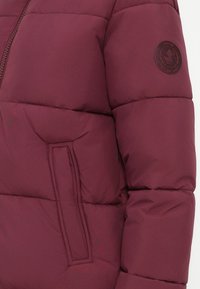 Jachetă puffer burgund cu un design matlasat, având un buzunar pe mânecă și o patch cu logo rotund pe partea superioară a brațului.