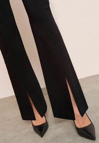Čierne rozšírené nohavice so bočnými rozparkami, v kombinácii s čiernymi špicatými textúrovanými lodičkami. Hladká tkanina s čistým dizajnom.