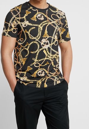 Czarny t-shirt z krótkim rękawem z wzorem złotych łańcuchów i czaszek, ozdobiony frędzlami. Materiał wydaje się gładki i lekki.