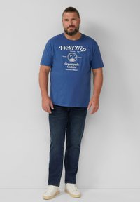 Mężczyzna z brodą, noszący niebieską koszulkę "Field Trip Countryside Culture", ciemne dżinsy i białe trampki, stojący na tle jasnej, gładkiej ściany.