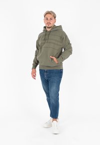 Felpa verde oliva con righe orizzontali, tasca frontale e cappuccio con cordino, abbinata a jeans blu e sneakers bianche su uno sfondo bianco.