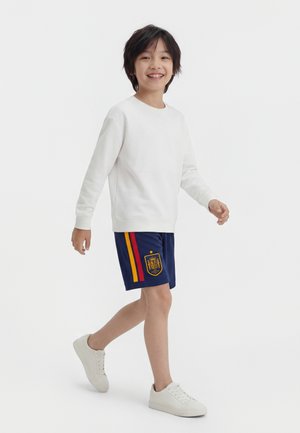 Παιδί που περπατά φορώντας λευκό φούτερ, σκούρο μπλε σορτς με κόκκινες και κίτρινες ρίγες και έμβλημα, καθώς και λευκά αθλητικά παπούτσια σε λευκό φόντο.