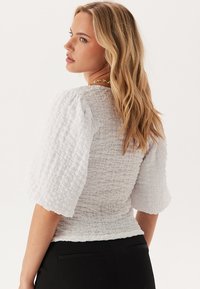 Top bianco con texture, maniche a sbuffo e design arricciato. Presenta una vita aderente e un tessuto morbido e leggero per il massimo comfort.