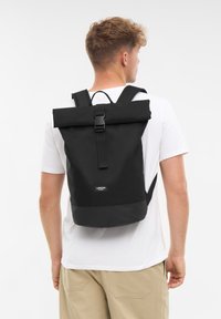 Férfi fekete, hátul csattal ellátott roll-top hátizsákot visel, fehér pólóval és bézs nadrággal egy egyszínű világos háttér előtt.