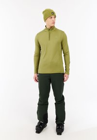 Man in een olijfgroene beanie, bijpassend ritsjack, donkergroene broek en zwarte skiboots staat tegen een witte achtergrond.