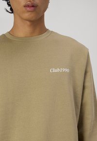 Khaki sweatshirt van zachte stof. Heeft een ronde hals en tekst op de linkerborst die "Club1990" in het wit zegt. Eenvoudig ontwerp.