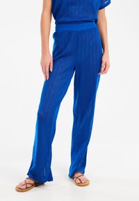 Blauwe gebreide wijde broek met een diamantpatroon, voorzien van een brede elastische tailleband en gecombineerd met bruine slippers.