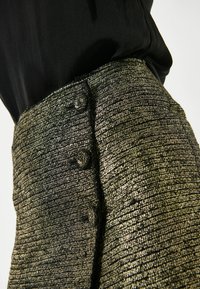 Tekstūruotas metalizuotas auksinis ir juodas sijonas su prigludusia juosmeniu, dekoratyviniais mygtukais šone ir lygiu audiniu.