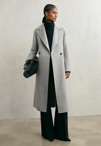 Svetlo sivi volneni plašč z zarezanim ovratnikom in dvojno zapenjanjem na gumbe, kombiniran s črnim puloverjem z visokim ovratnikom in širokimi hlačami. Črn modni dodatki v obliki torbice.