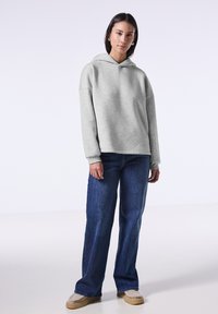 Sudadera gris clara con un acabado texturizado, que presenta una capucha con cordón, hombros caídos y un corte relajado, combinada con unos pantalones vaqueros de pierna ancha azul oscuro.