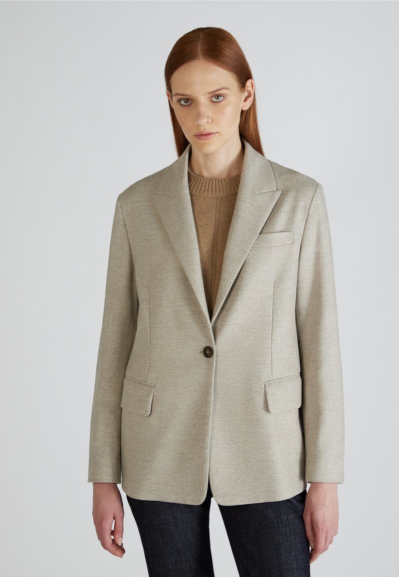 Blazer su misura beige chiaro con un singolo bottone, colletto a rever e due tasche frontali. Tessuto strutturato, abbinato a un maglione marrone sotto.