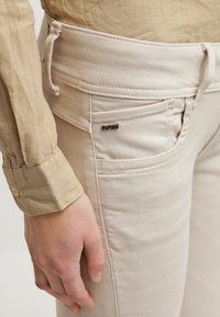 Pantalones de algodón beige con corte slim, que cuentan con bolsillos frontales y una pequeña etiqueta negra en el lateral. Mano reposando en la cadera.