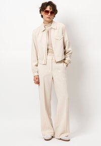 Beige cropped jack met een puntige kraag en elastische manchetten, gedragen met beige wijde broekspijpen. Lichte getextureerde coltrui eronder. Bril inbegrepen.