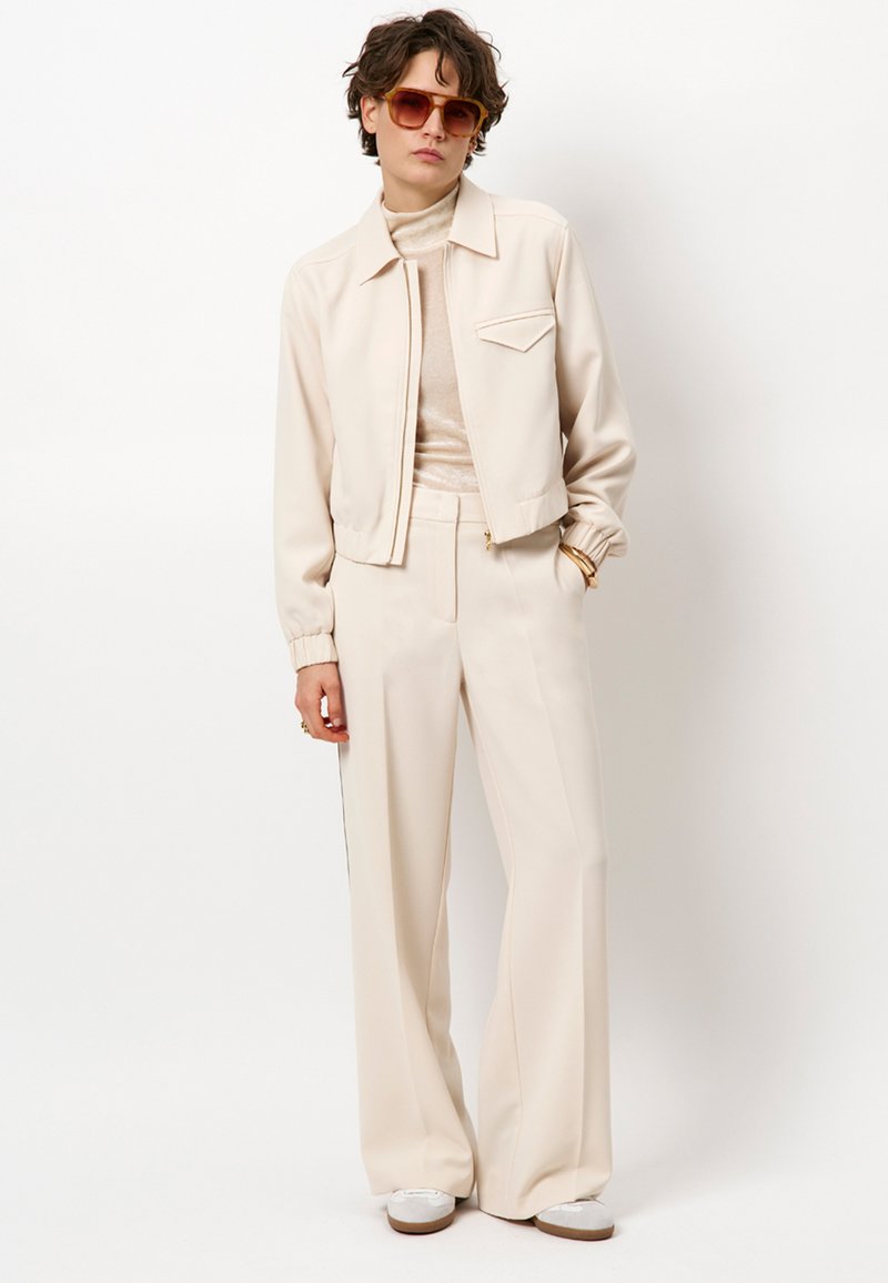 Beige cropped jack met een puntige kraag en elastische manchetten, gedragen met beige wijde broekspijpen. Lichte getextureerde coltrui eronder. Bril inbegrepen.