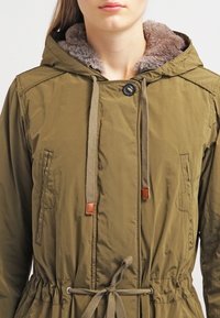 Mujer con parka verde oliva con capucha y forro de piel, cordones en la capucha y la cintura, y bolsillos frontales en ángulo.