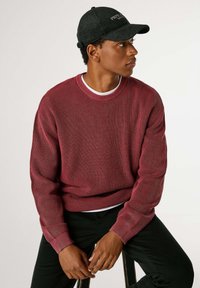 Roter strukturiert gestrickter Pullover mit rundem Ausschnitt und gerippten Bündchen, über einem weißen Hemd getragen, kombiniert mit schwarzen Hosen.