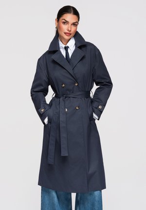 Donna che indossa un trench doppiopetto blu navy con cintura, camicia bianca, cravatta nera e jeans blu a gamba larga, mani nelle tasche del cappotto.