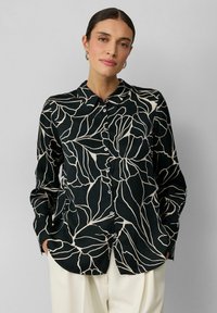 Zwarte overhem shirt met wit bladdessin, featuring een kraag en lange mouwen, gemaakt van een gladde stof. Gdragen met crèmekleurige broek.
