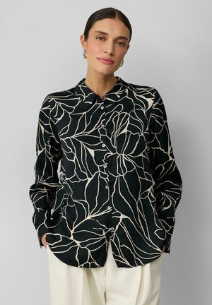 Camicia nera con bottone, decorata con un motivo di foglie bianche, dotata di colletto e maniche lunghe, realizzata in tessuto morbido. Indossata con pantaloni color crema.