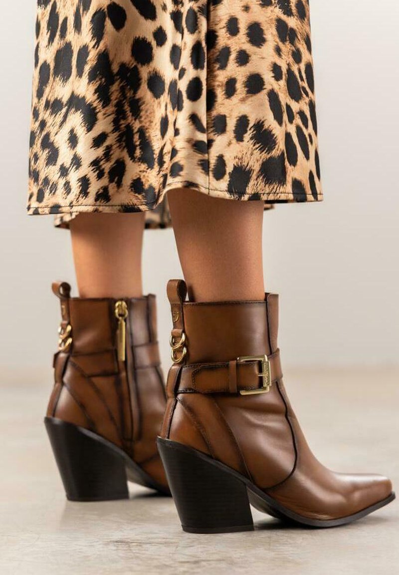 Botas de tobillo de cuero marrón con punta cuadrada, un cinturón dorado y acentos de hardware, descansando sobre una falda de estampado de leopardo.