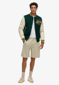 Giacca varsity verde e crema con strisce gialle, logo ricamato, indossata sopra una maglietta bianca e pantaloni corti beige. Sneakers beige e calzini.