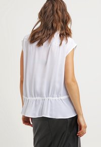 Weiße Bluse mit kurzen Ärmeln, die eine geraffte Taille und gestickte Details an der Rückseite aufweist, aus leichtem, fließendem Stoff gefertigt.