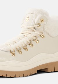 Biały skórzany but trekkingowy z futrzanym wykończeniem, złotymi oczkami i wiązaniem na sznurowadła. Teksturowana gumowa podeszwa z rowkami dla lepszej przyczepności.