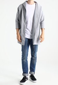 Light gray, oversized cardigan with a hood, crafted from soft fabric. Worn over a white shirt, paired with blue jeans and black sneakers.