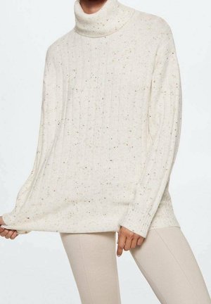 Personne portant un pull à col roulé crème tacheté et un pantalon beige clair, tenant l'ourlet du pull d'une main.