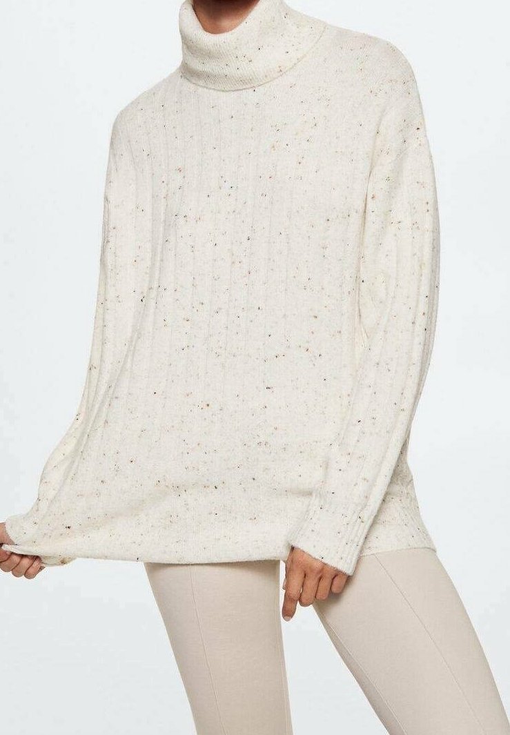Personne portant un pull à col roulé crème tacheté et un pantalon beige clair, tenant l'ourlet du pull d'une main.